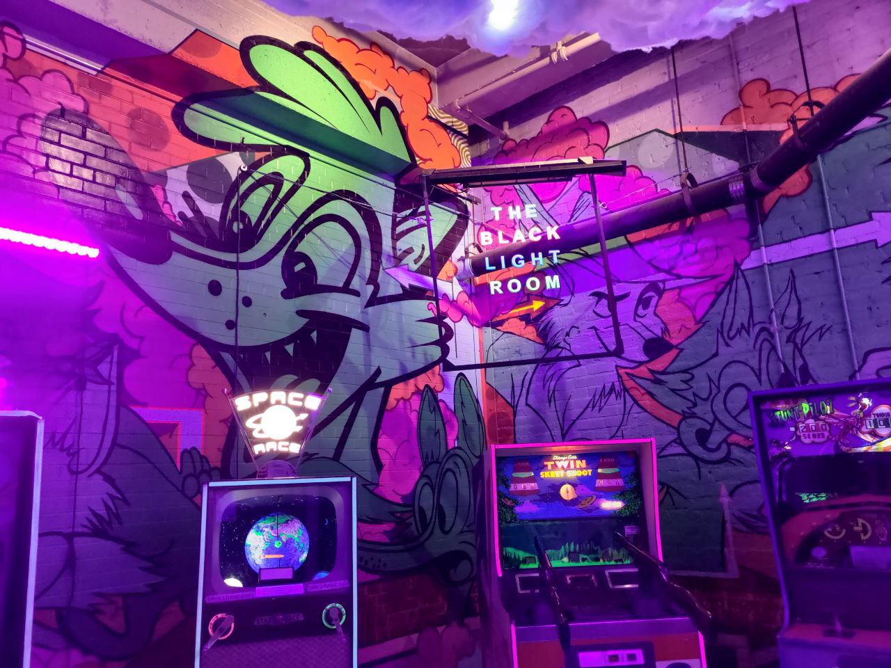 A photo from the back room of Can Can Wonderland, called The Black Light Room. The only light in this room is from blacklight bulbs. Arcade machines stand along the wall, which has been painted various shades of purples, greens, and oranges. The wall is a floor-to-ceiling mural of differerent cartoon foxes, including Disney's Robin Hood
