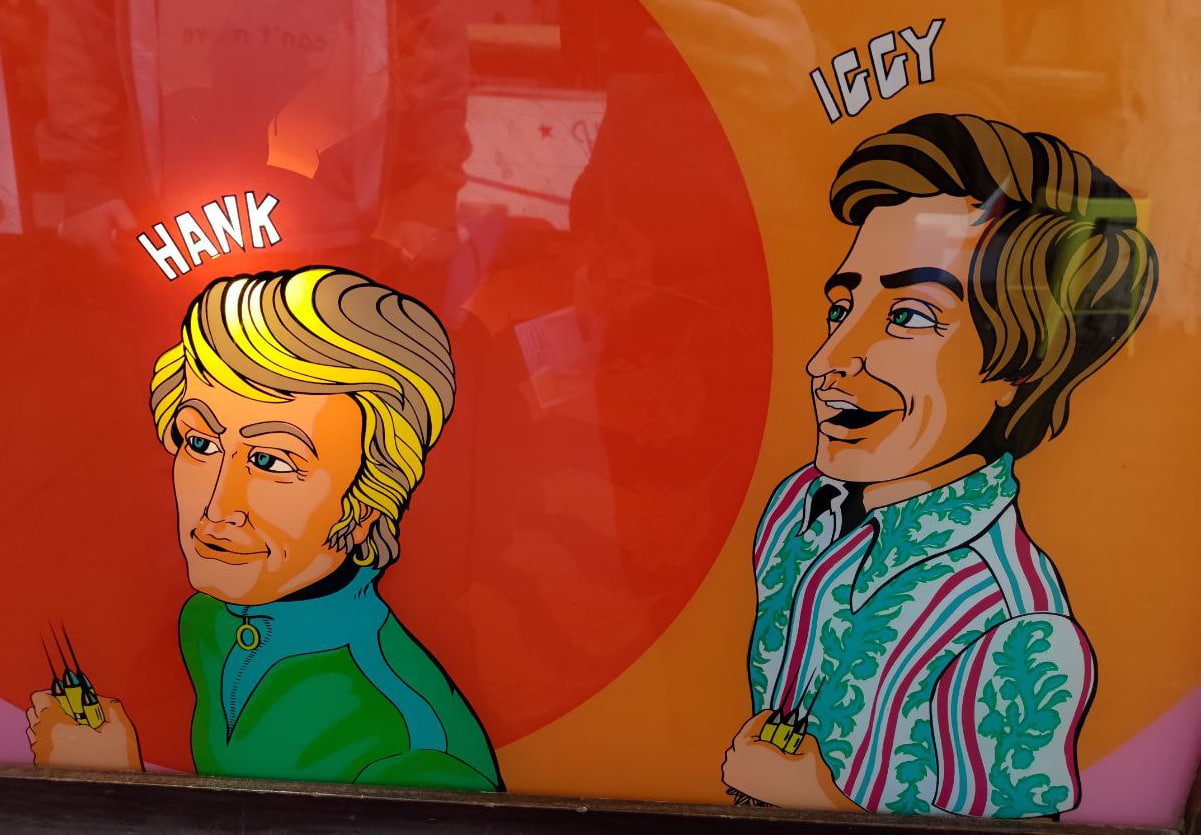A photo of the art from Bull's Eye. Two guys, one named Hank and one named Iggy, stand there clutching a fistful of darts. Hank has dirty blonde hair and is wearing a green and blue jacket. Iggy has brown hair and is wearing some sort of stripe and floral print button up.